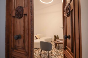 Uma porta de madeira com maçaneta entalhada, um sofá branco com almofada estampada, uma cadeira cinza e uma pequena mesa com um vaso.