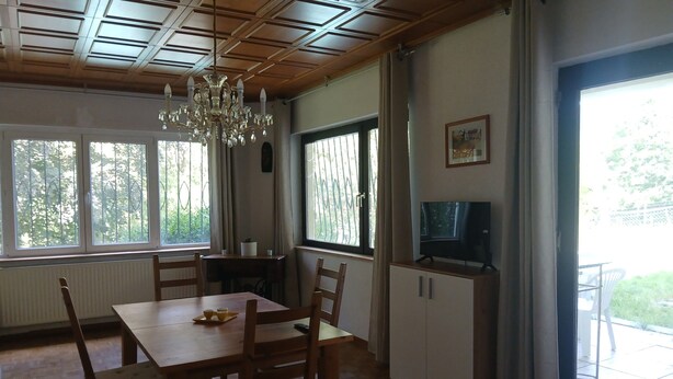 A dining area with a wooden table and chairs, a chandelier, a television on a cabinet, and large windows with curtains. - Apartment 'Weidenblick' with Mountain View, Private Terrace and Wi-Fi (Schottwien)