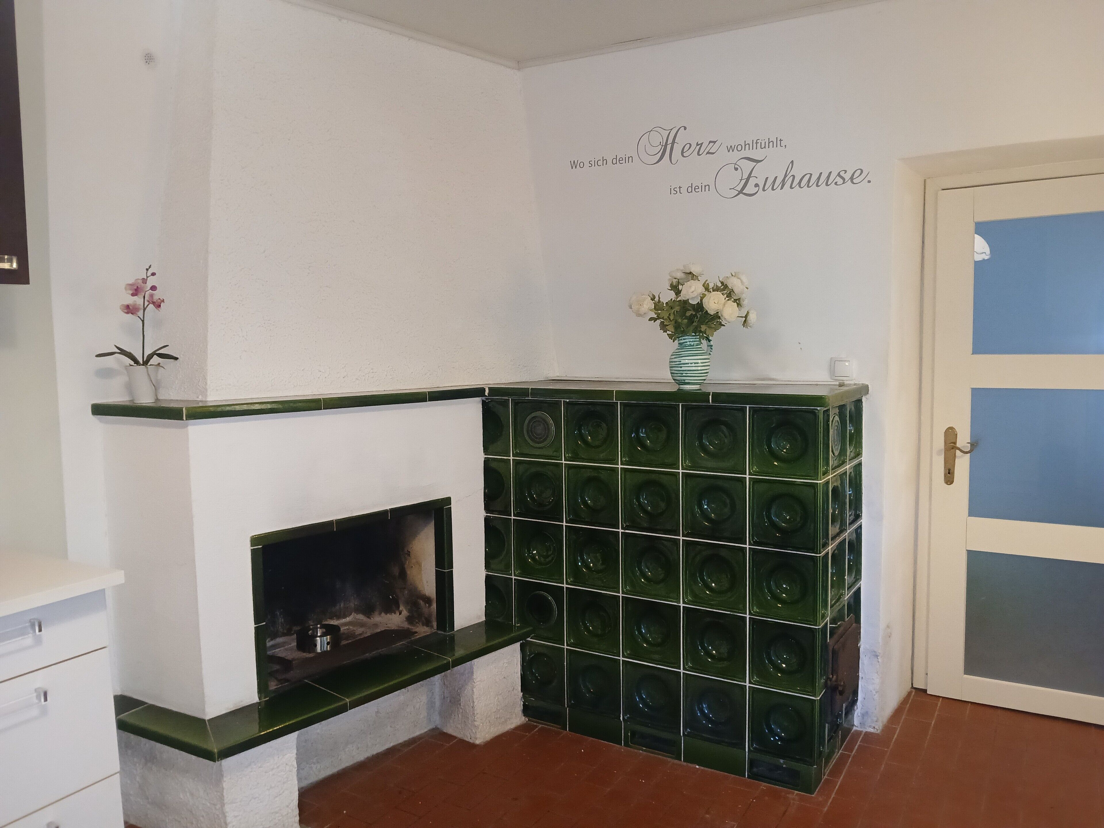 A room with a fireplace, a wall with a decorative phrase, a green tiled structure, and a door with a blue panel.