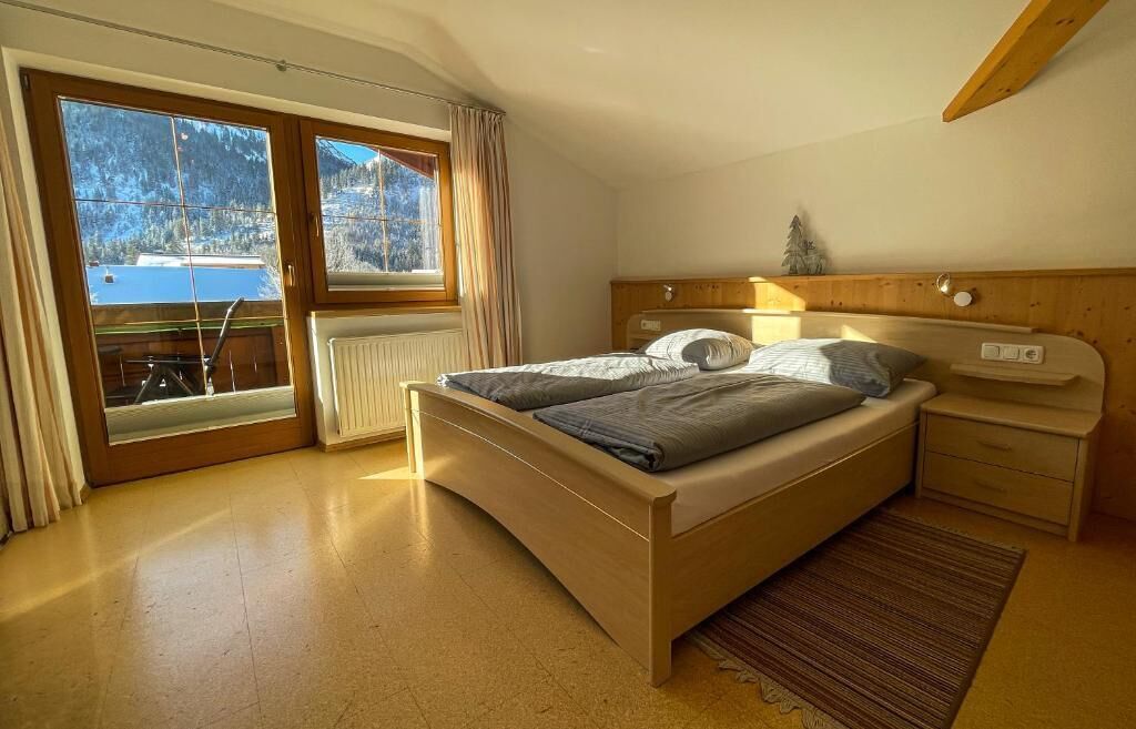 Un dormitorio con cama, mesita de noche, ventana con cortinas y vista a un paisaje montañoso.