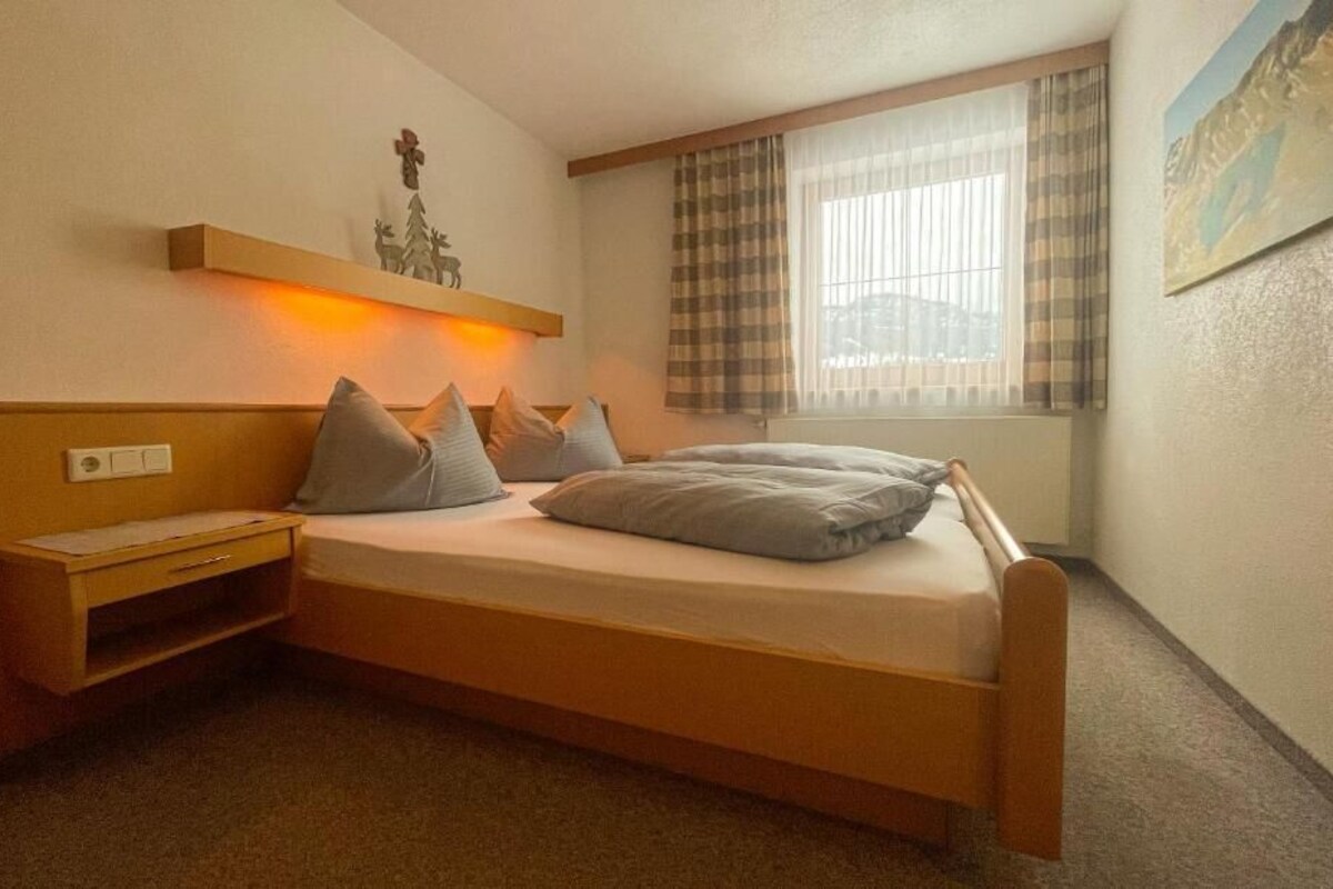 A single bed with a headboard, a bedside table, and a nightstand. There is a window with curtains, a wall-mounted shelf with a small decoration, and a painting on the wall.