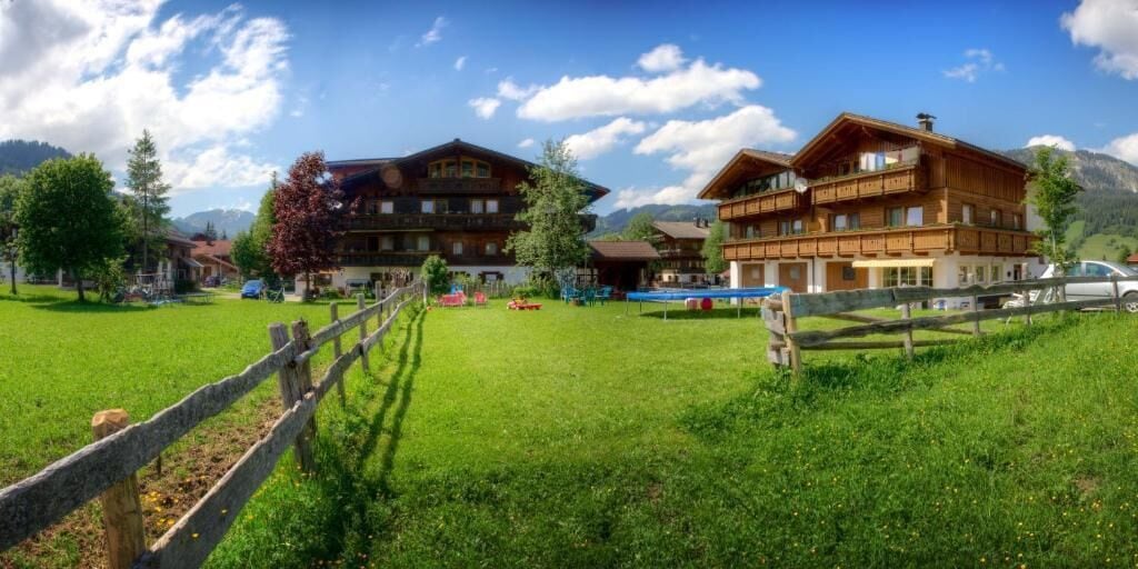 Ein Chalet-ähnliches Holzhaus mit Balkon, umgeben von einer Wiese und Bergen im Hintergrund.