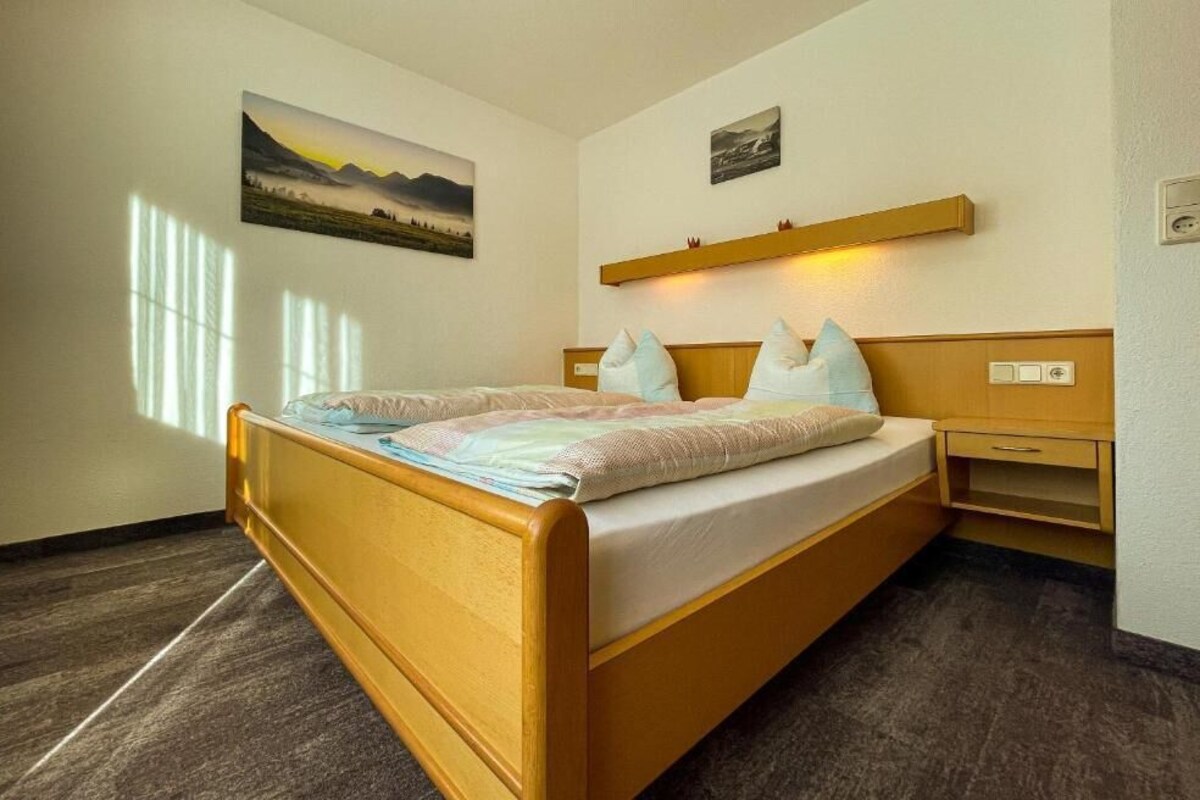 A double bed with a wooden frame and a headboard, a bedside table with a lamp, and a wall-mounted shelf with a picture.