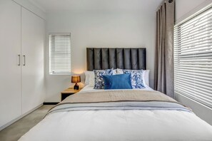 Ein Schlafzimmer mit Bett, Nachttisch, Lampe und Jalousien.