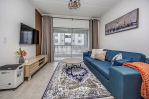Ein modernes Wohnzimmer mit einem blauen Sofa, einem kleinen runden Tisch, einem an der Wand montierten Fernseher und einem großen Fenster mit Jalousien.
