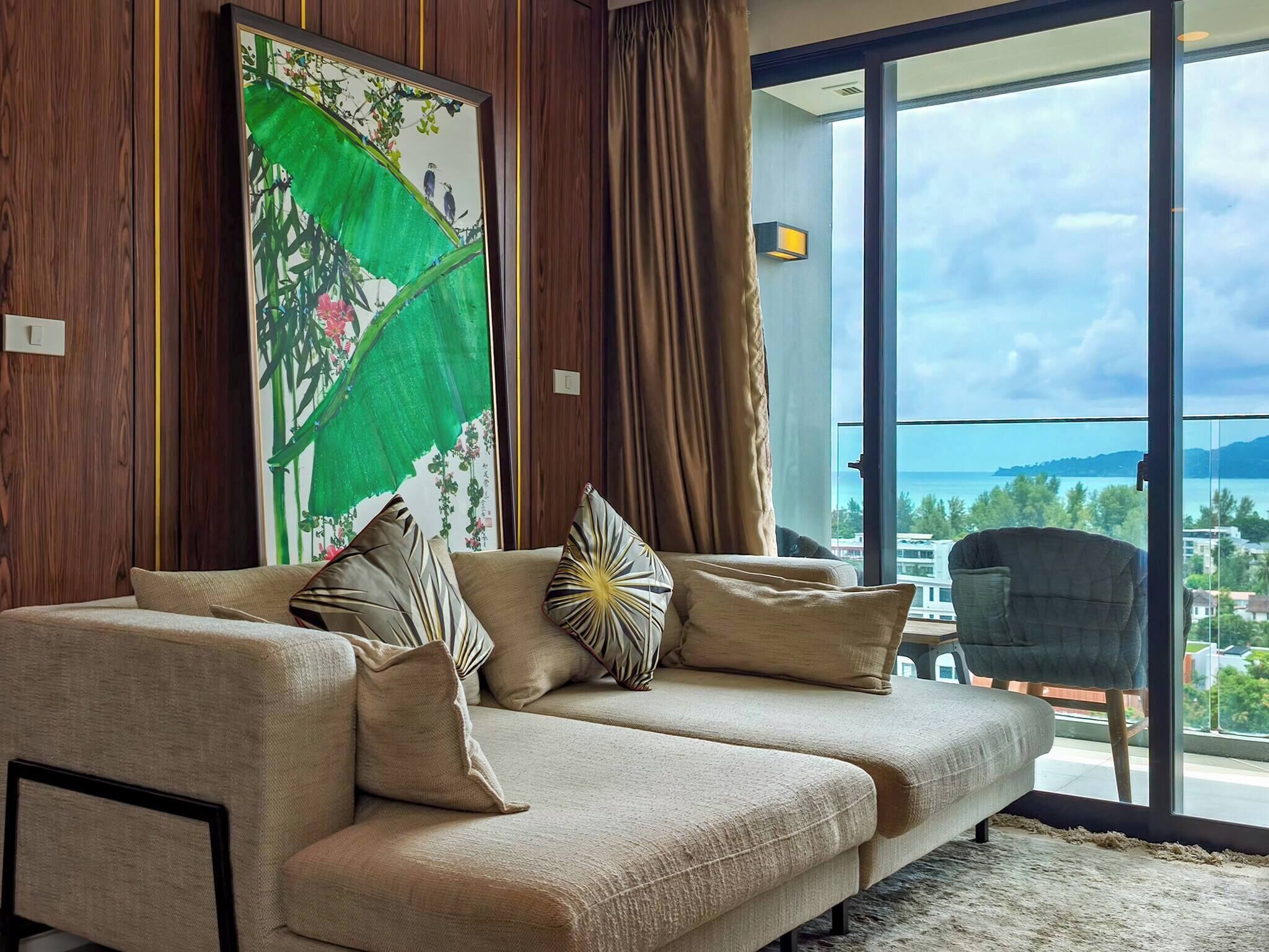 A modern living room with a sofa, decorative pillows, a large window with a view, and a painting on the wall.