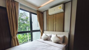 A hotel room with a large bed, a window with curtains, an air conditioning unit, and a view of greenery outside. - Luxurious 2BR Condo with Ocean Views in Choeng Thale (Tambon Choeng Thale)