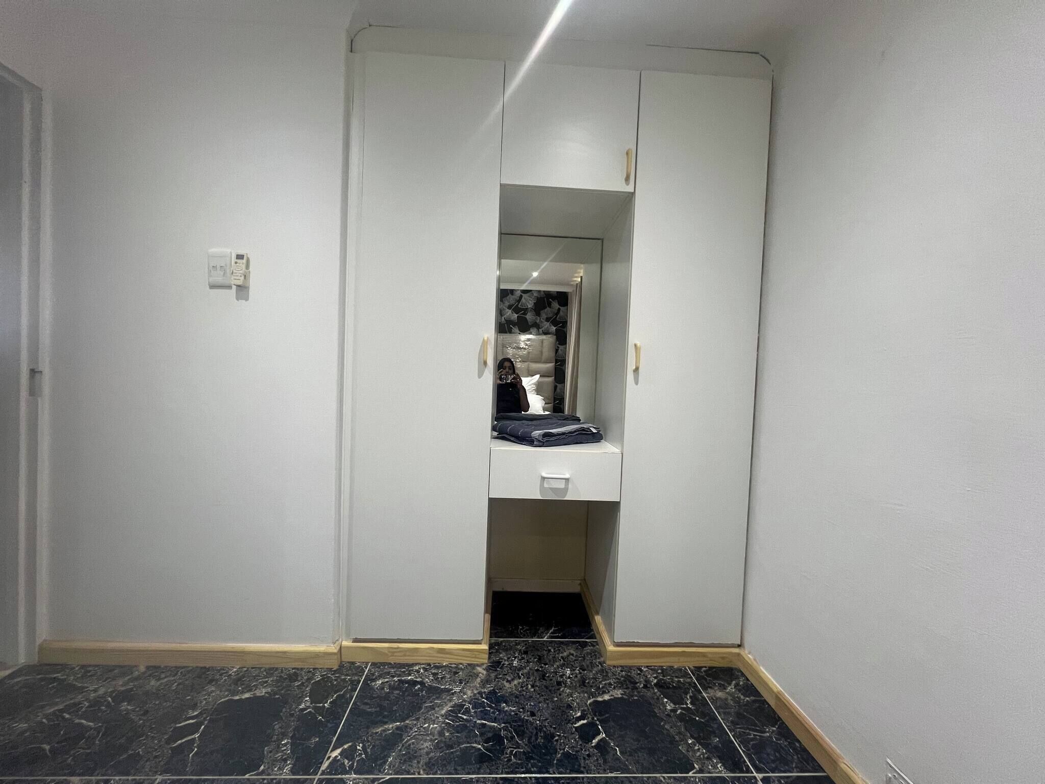 Un pasillo estrecho con una puerta de armario espejada, un mueble blanco pequeño y un suelo de baldosas negras.