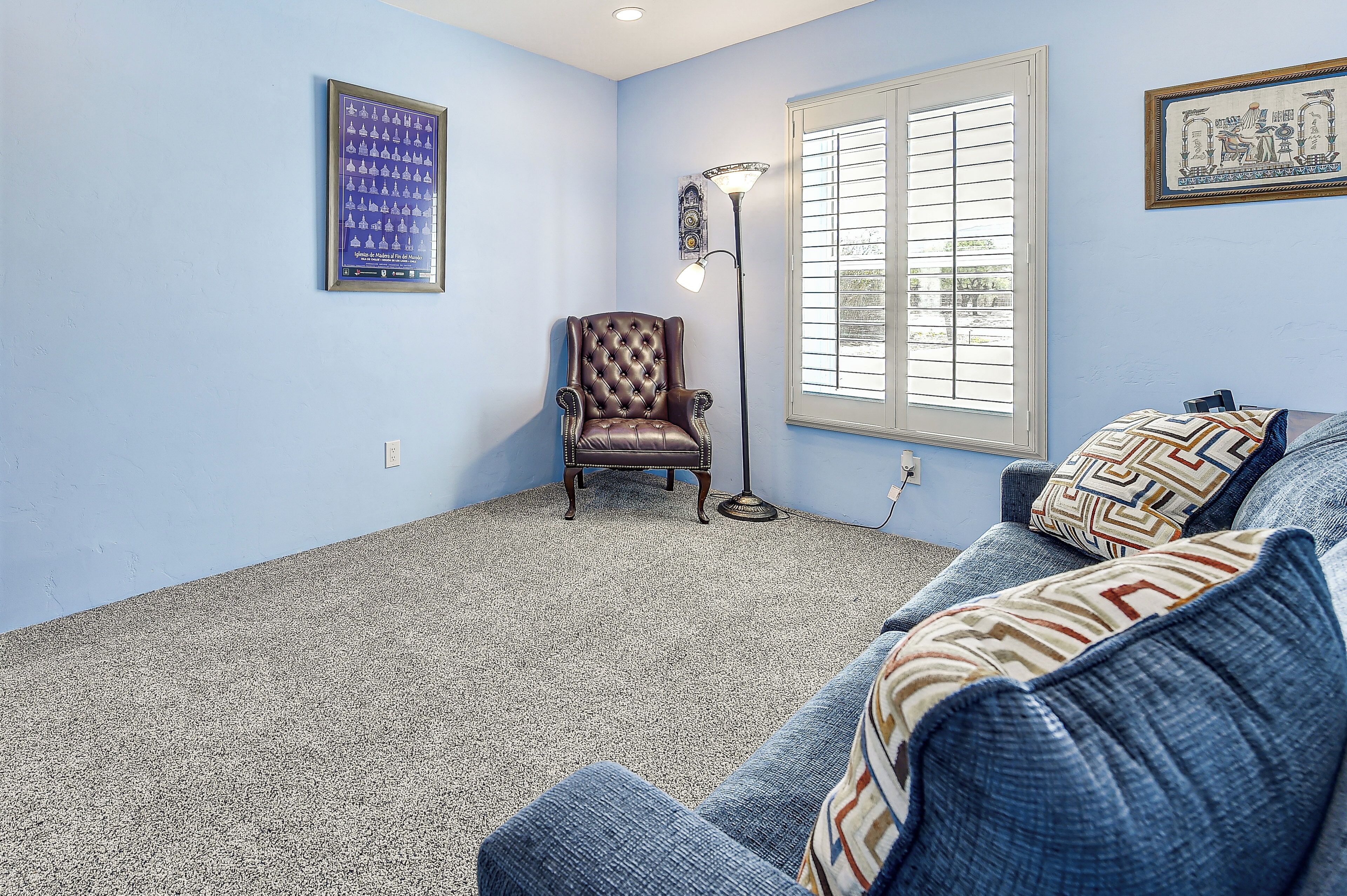 Quarto com parede azul, carpete estampado, poltrona de couro marrom, sofá azul com almofadas estampadas e janela com persianas.