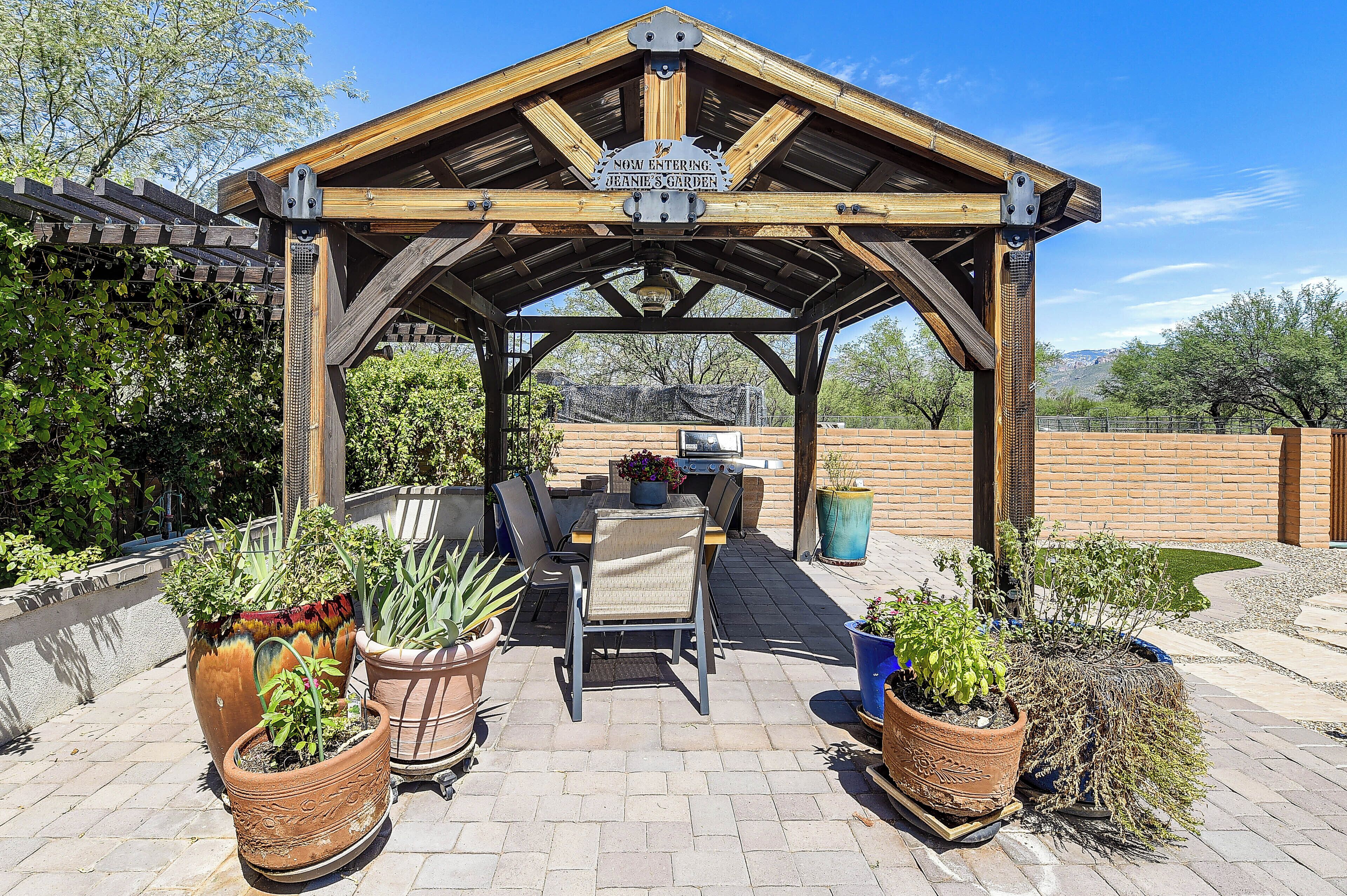Une pergola en bois sur laquelle est accrochée une pancarte indiquant « Vous entrez maintenant dans le jardin de Jeanie », ainsi qu’un espace patio aménagé avec des chaises et des plantes en pot.