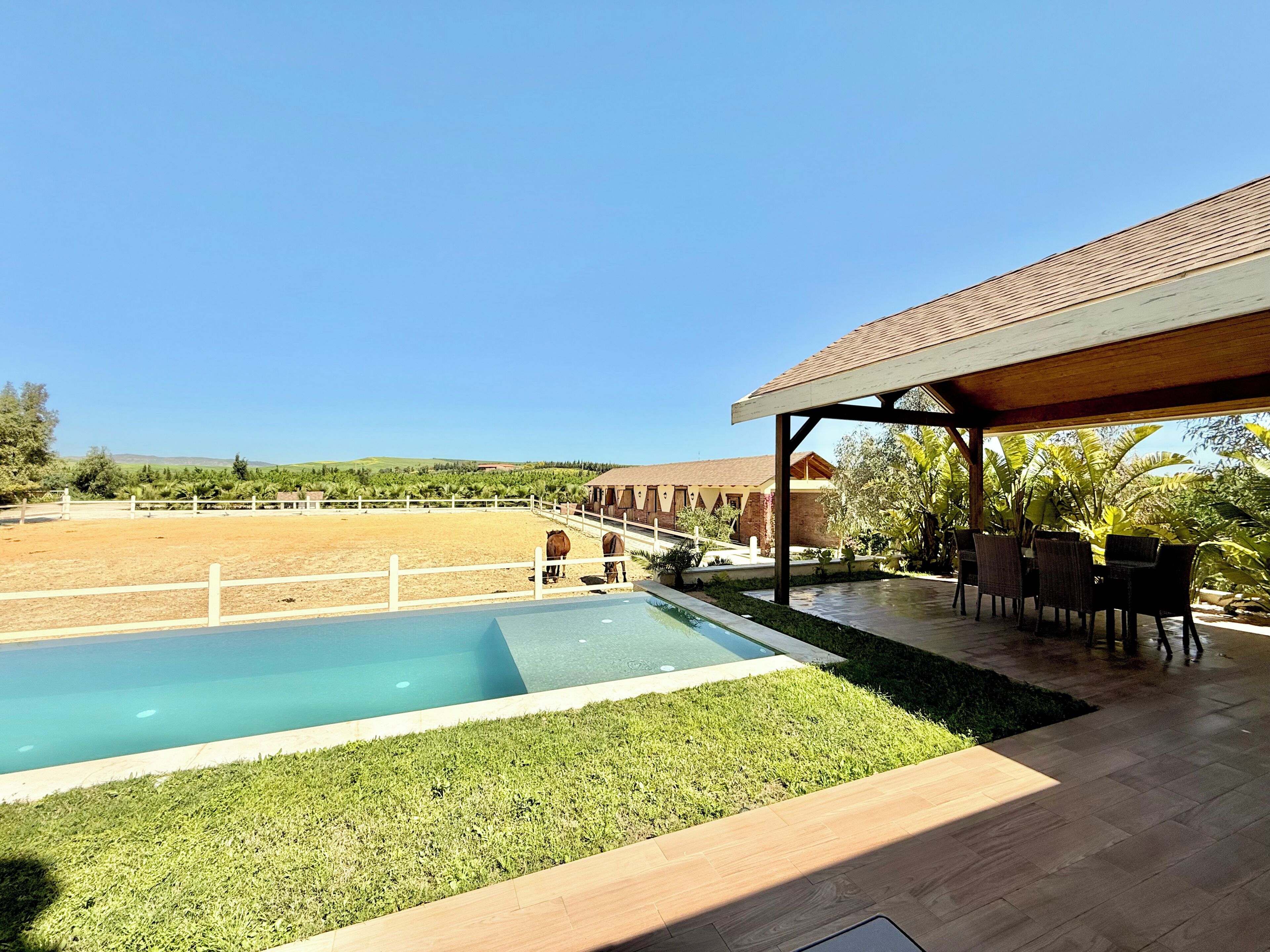 Una piscina con césped, un porche cubierto con sillas y un corral cercado.