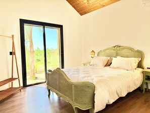 Quarto com cama, mesa de cabeceira, cadeira e janela com vista para o verde.
