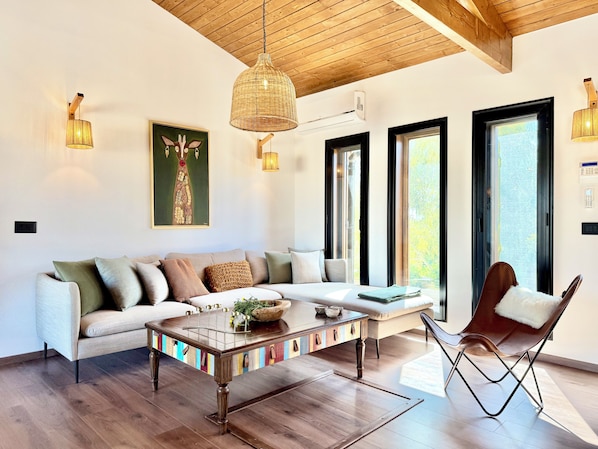 Uma sala de estar moderna com sofá, mesa de centro e uma poltrona. Há janelas amplas e um teto com vigas de madeira.