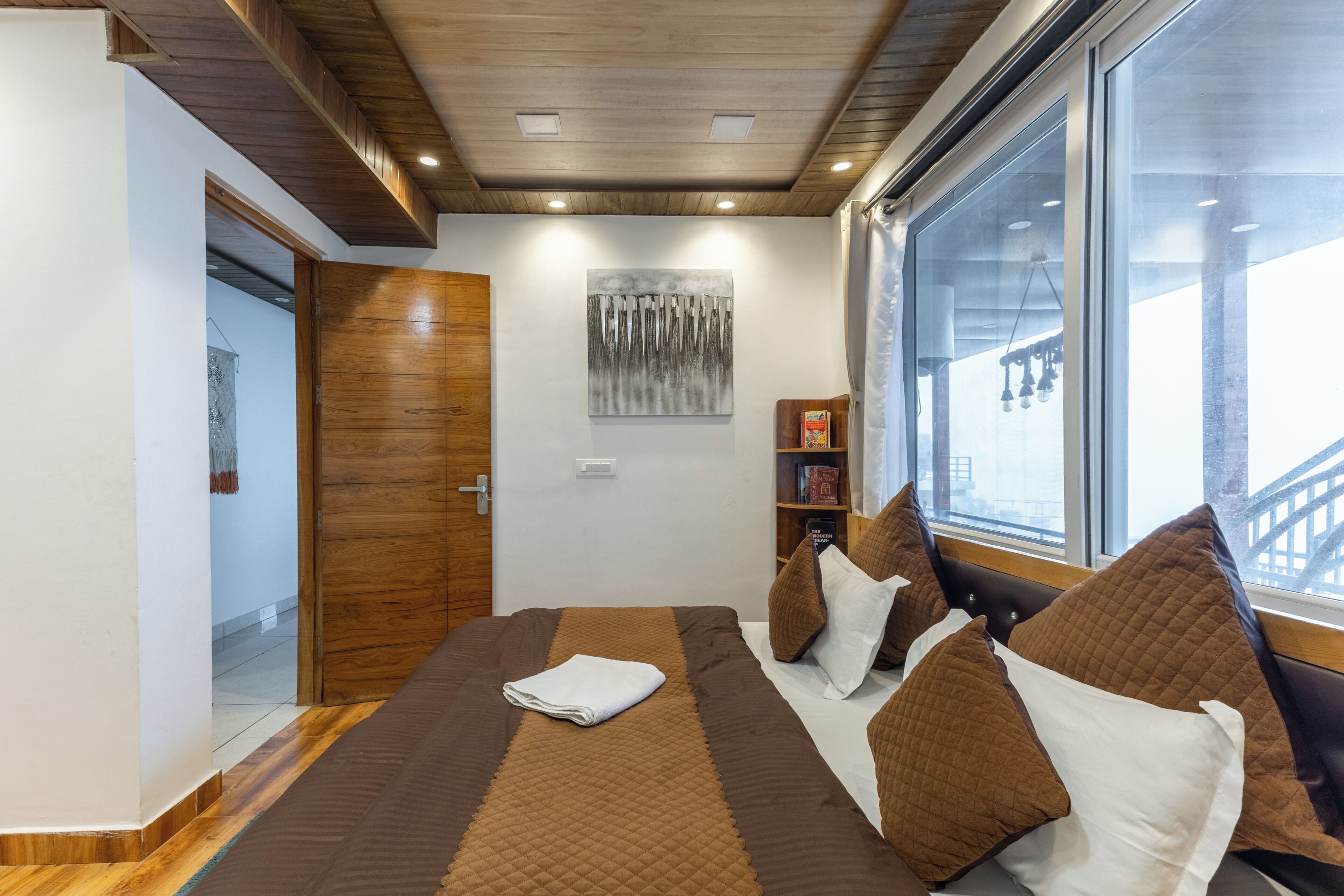 Una habitación de hotel con cama, ventana con vistas, puerta de madera y una obra de arte colgada en la pared.