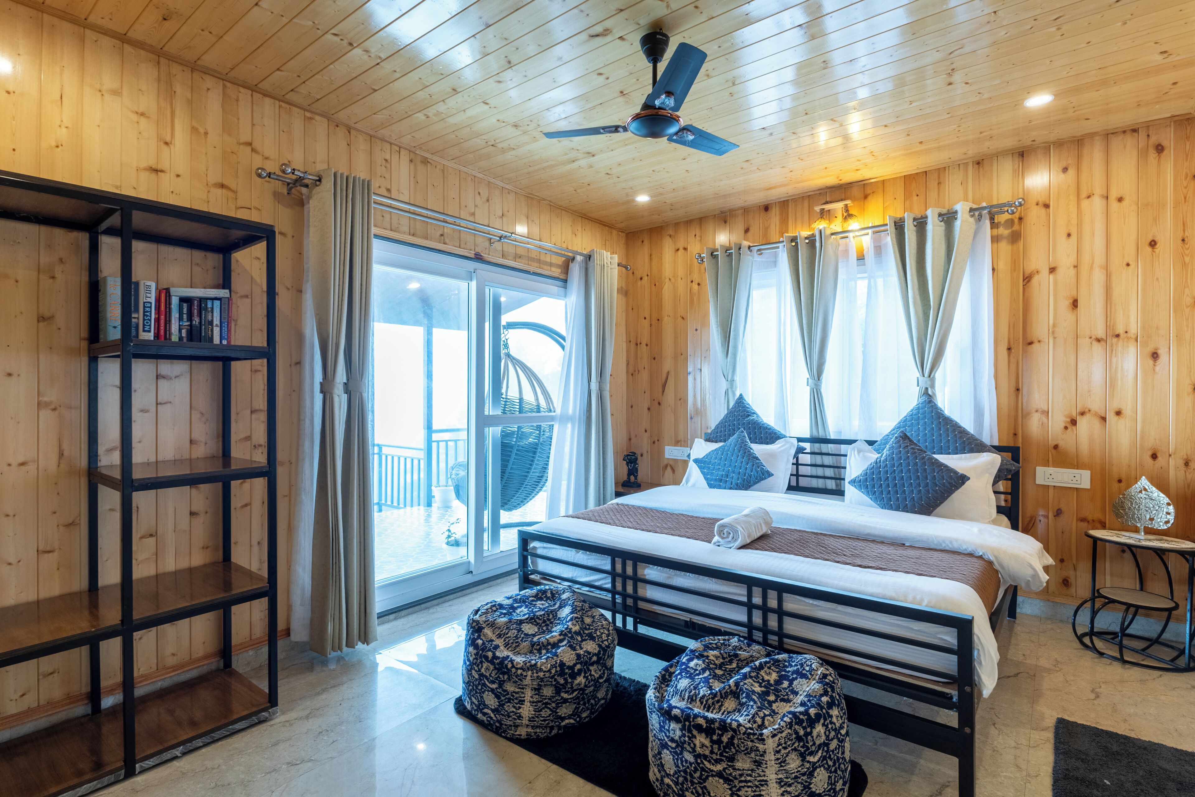 A wooden-paneled bedroom with a bed, two ottomans, a bookshelf, and a balcony view.