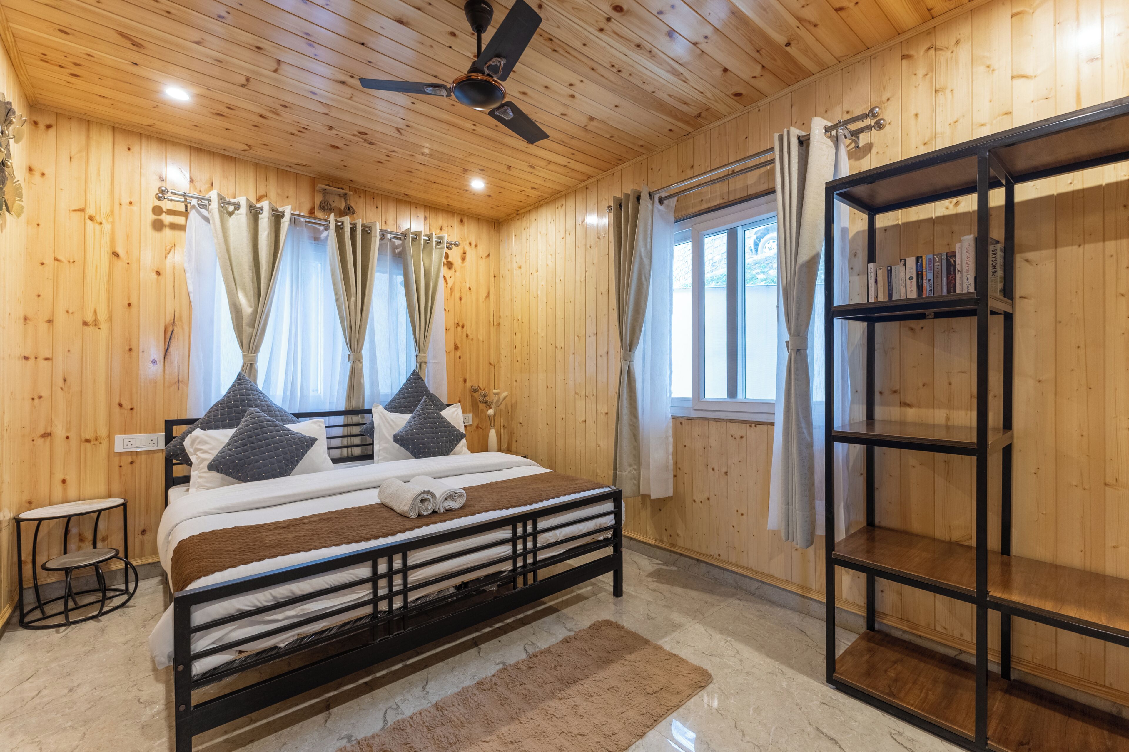 Un dormitorio con paneles de madera, una cama, un ventilador de techo, una ventana con cortinas y una estantería.