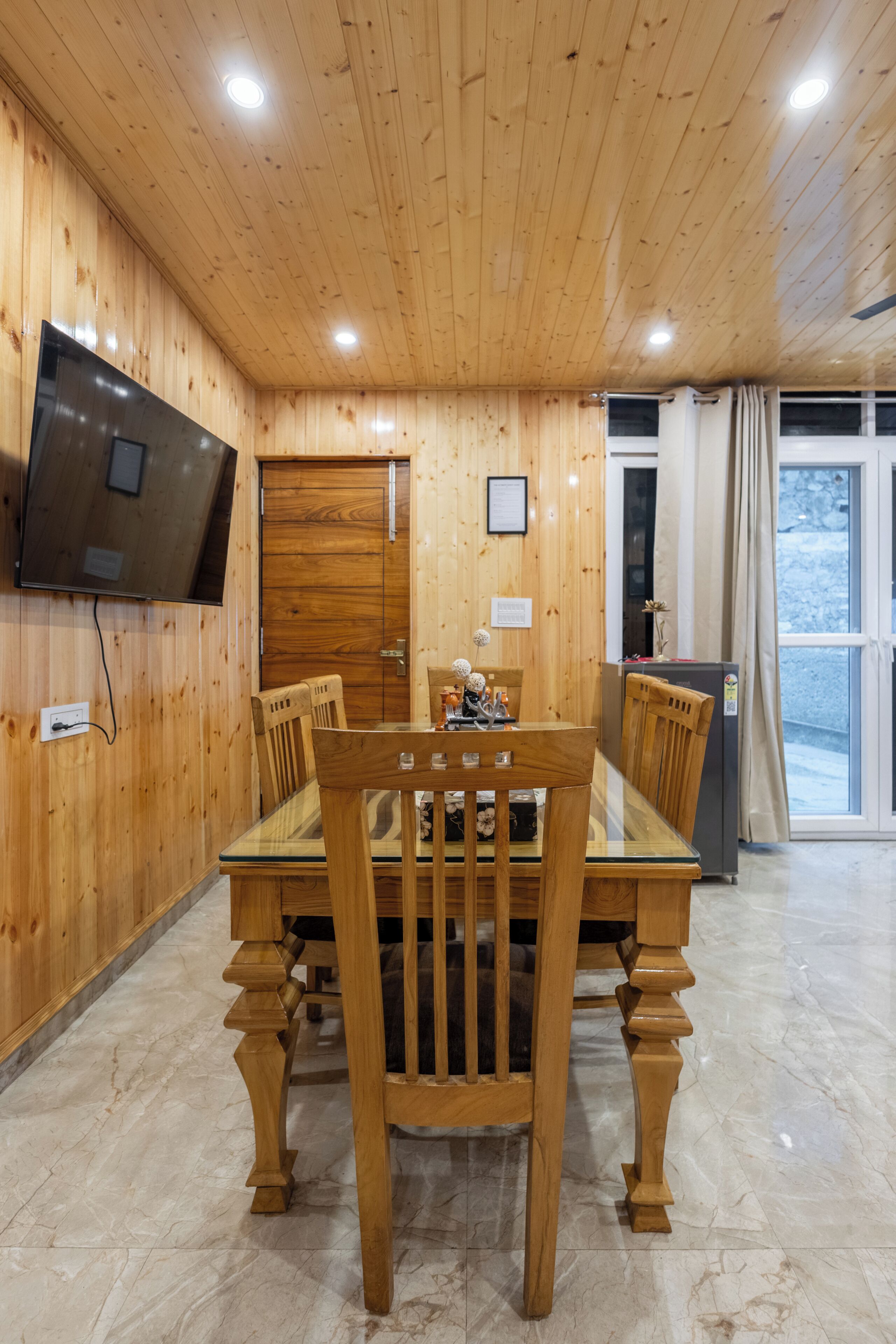Una habitación con paneles de madera, una mesa de comedor, sillas, un televisor montado en la pared y una puerta que da al exterior.