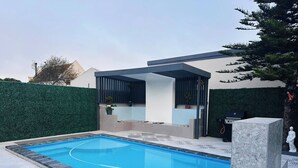Una piscina con un área de estar cubierta, un muro de piedra y un seto.