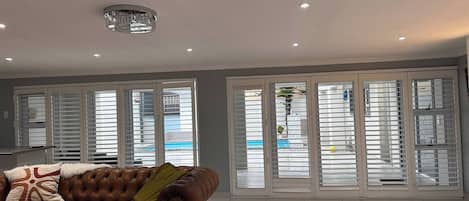 A spacious living room with a brown leather sofa, white tiled floor, and large glass doors with white blinds.