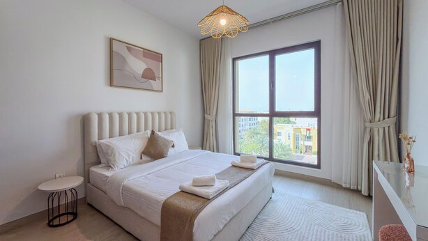 A hotel room with a large bed, a bedside table, a small round table, a window with curtains, and a hanging light fixture. - Jadeel Residences- MJL - Dubai (Dubai)