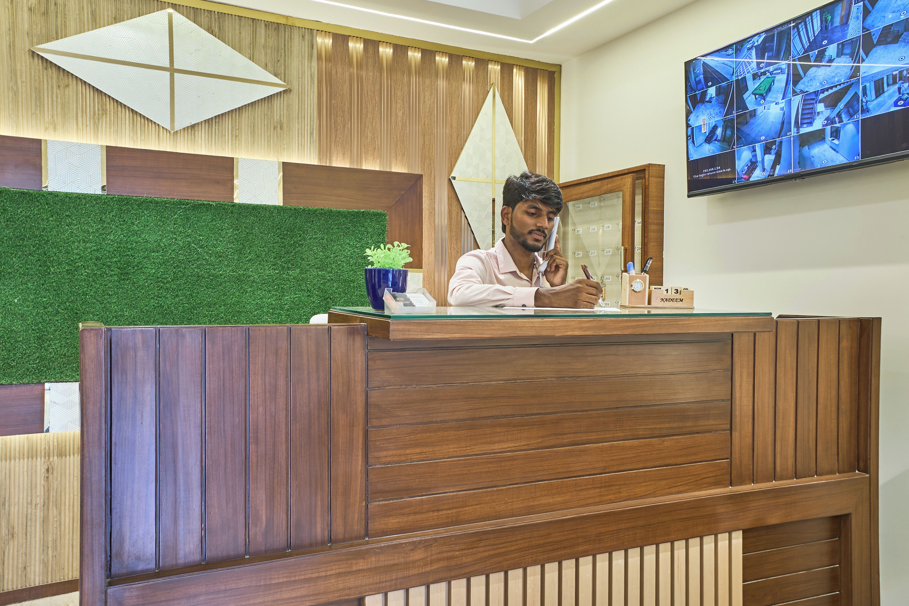Un mostrador de recepción con detalles de madera, un panel de pared con césped artificial verde y un monitor de cámara de seguridad en la pared.