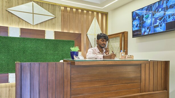 Un mostrador de recepción con detalles de madera, un panel de pared con césped artificial verde y un monitor de cámara de seguridad en la pared.