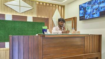 Un mostrador de recepción con detalles de madera, un panel de pared con césped artificial verde y un monitor de cámara de seguridad en la pared.