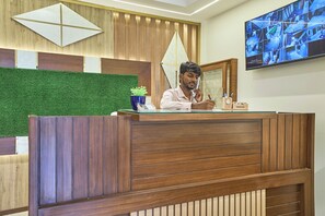 A reception desk with a wooden finish, a green artificial grass wall, and a security camera monitor on the wall. - Hotel Golden Place 325E (Gurugram)