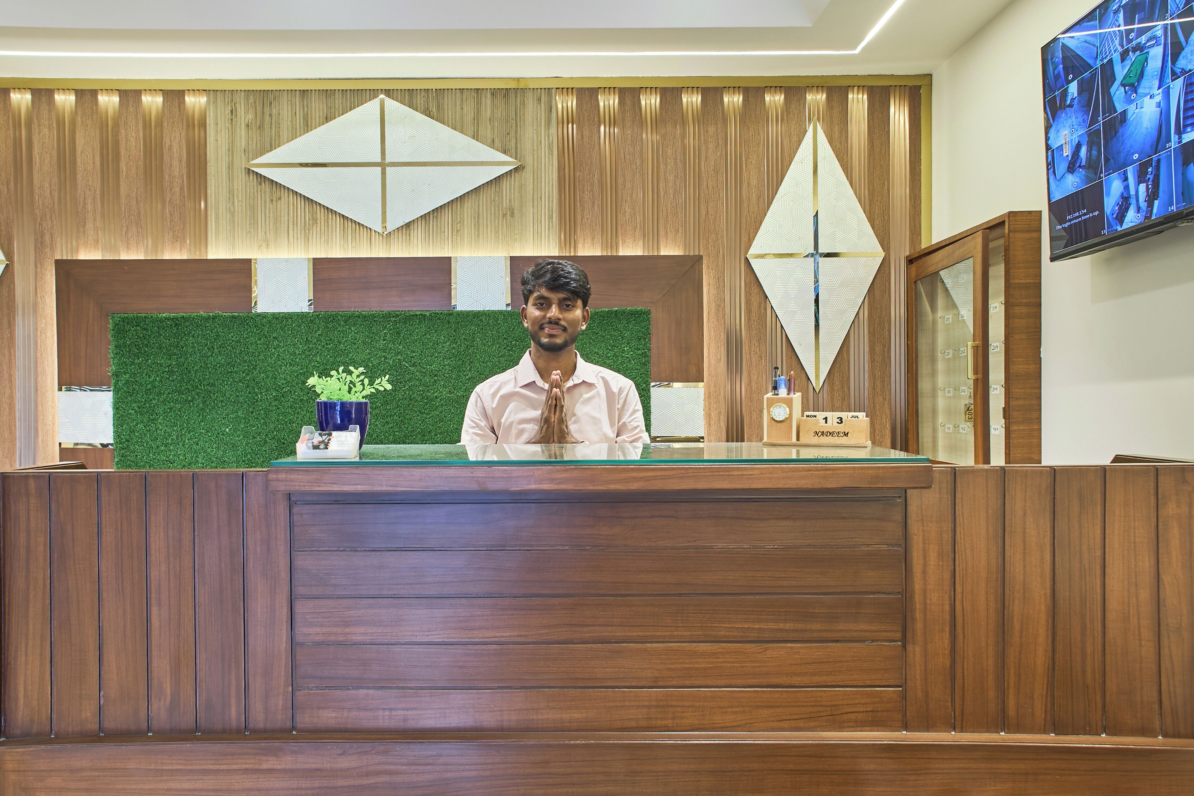 Un mostrador de recepción con detalles de madera, un fondo de césped artificial verde y un reloj digital mostrando la hora.