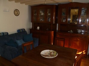 Ein Essbereich mit einem Holztisch und Stühlen, ein blaues Sofa mit Kissen sowie eine Küchenzeile mit Holzschränken und einer Spüle.