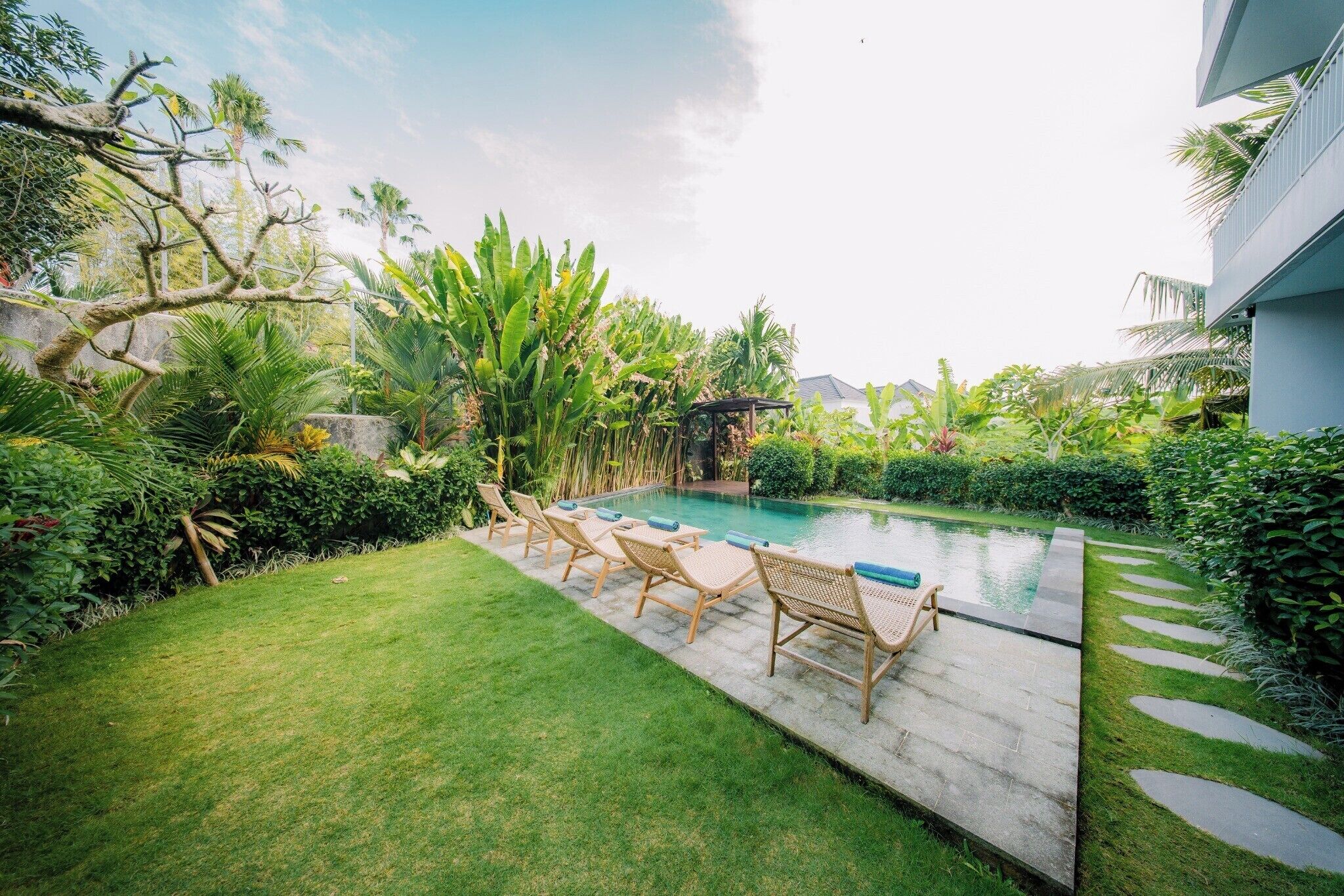 Un área de piscina con sillones reclinables, césped cuidado y vegetación tropical.