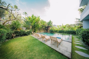 Un área de piscina con sillones reclinables, césped cuidado y vegetación tropical.