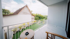 A balcony with a white railing, a small round table, and a chair. There is a view of a house with a tiled roof and a garden with potted plants. - Puspa Guest House (Pererenan)