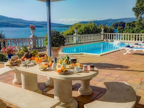A poolside breakfast setup with a table, fruits, and beverages, overlooking a body of water and hills. - Tolle Villa oberhalb der Bucht Ria de Vigo by Interhome (Pontevedra)