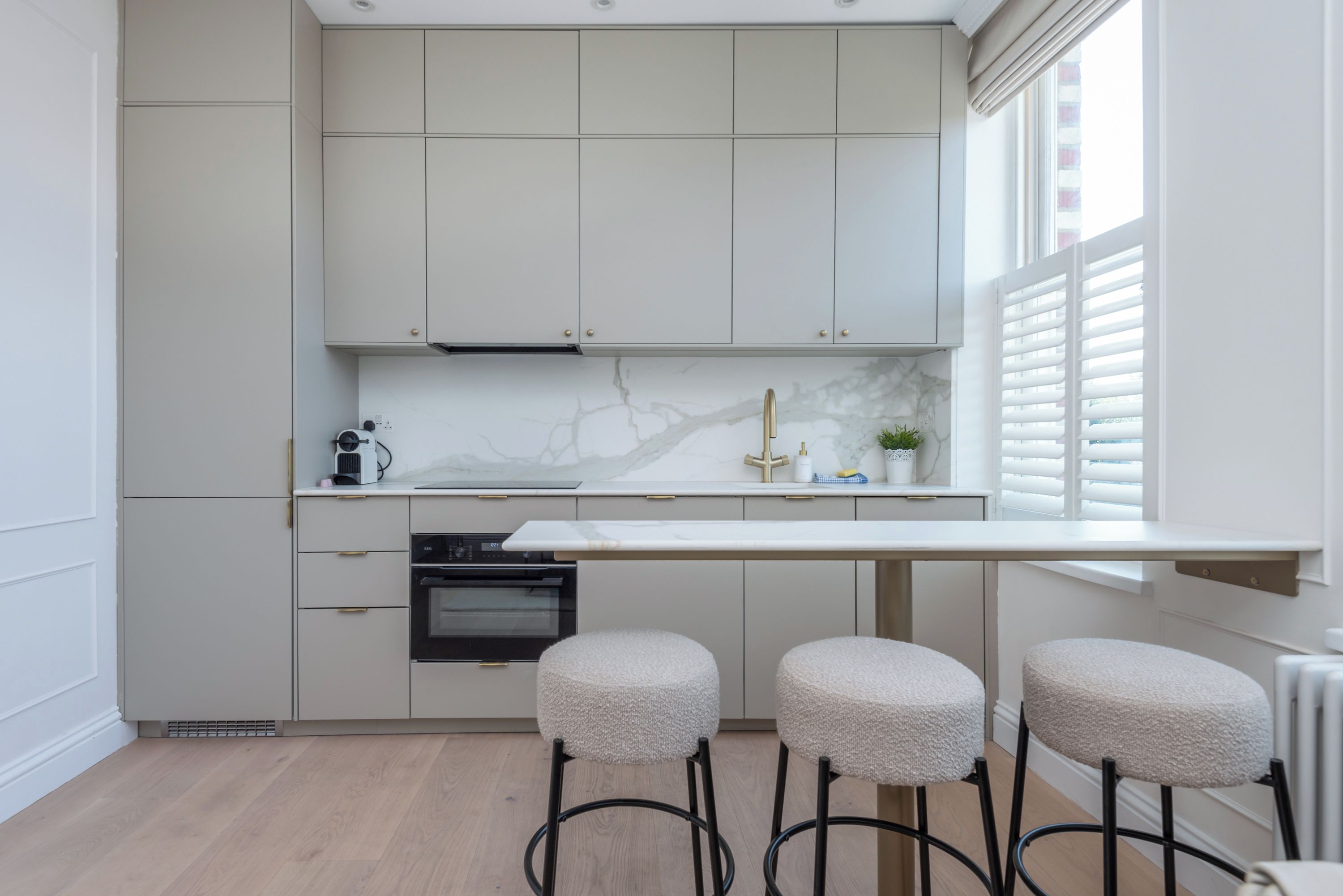 A modern kitchen with white cabinetry, a marble countertop, and a built-in oven. There are three stools and a window with blinds.