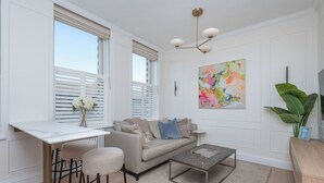 A modern living room with a sofa, coffee table, and bar stools. There is a window with blinds, a painting on the wall, and a plant. - Modern 2BDR with Workspace in Clapham Junction (Greater London)