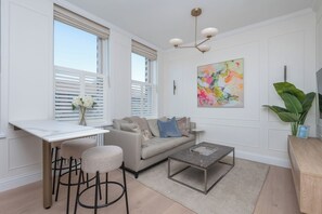 A modern living room with a sofa, coffee table, and bar stools. There is a window with blinds, a painting on the wall, and a plant. - Modern 2BDR with Workspace in Clapham Junction (Greater London)