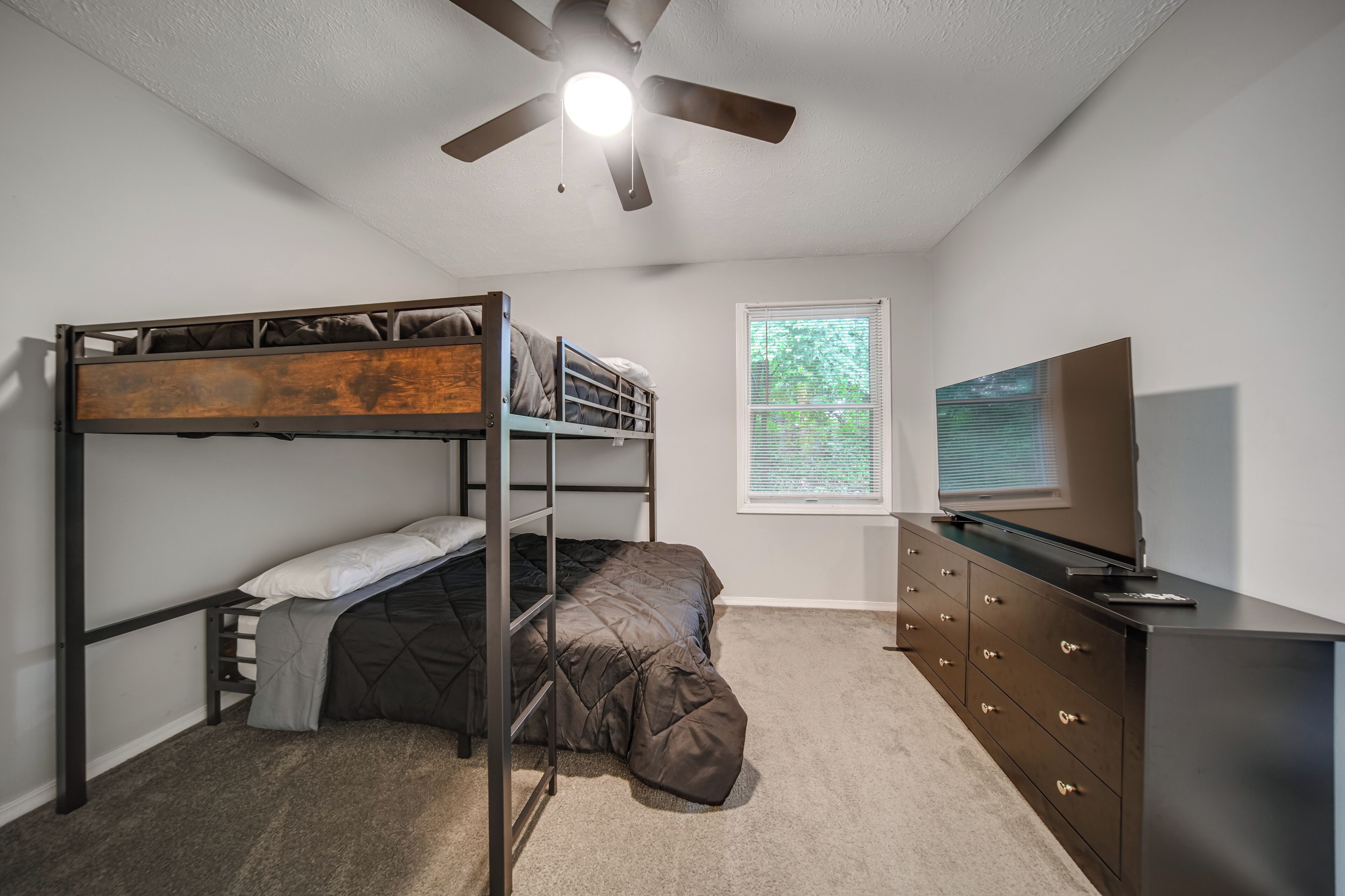 A bedroom with bunk beds, a ceiling fan, a window with blinds, a dresser, and a television.