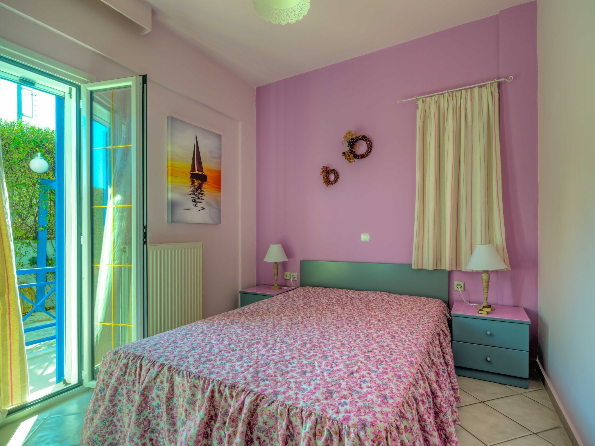 Un dormitorio con una cama, dos mesitas de noche, un ventanal con vistas y una pared con un cuadro y dos adornos.