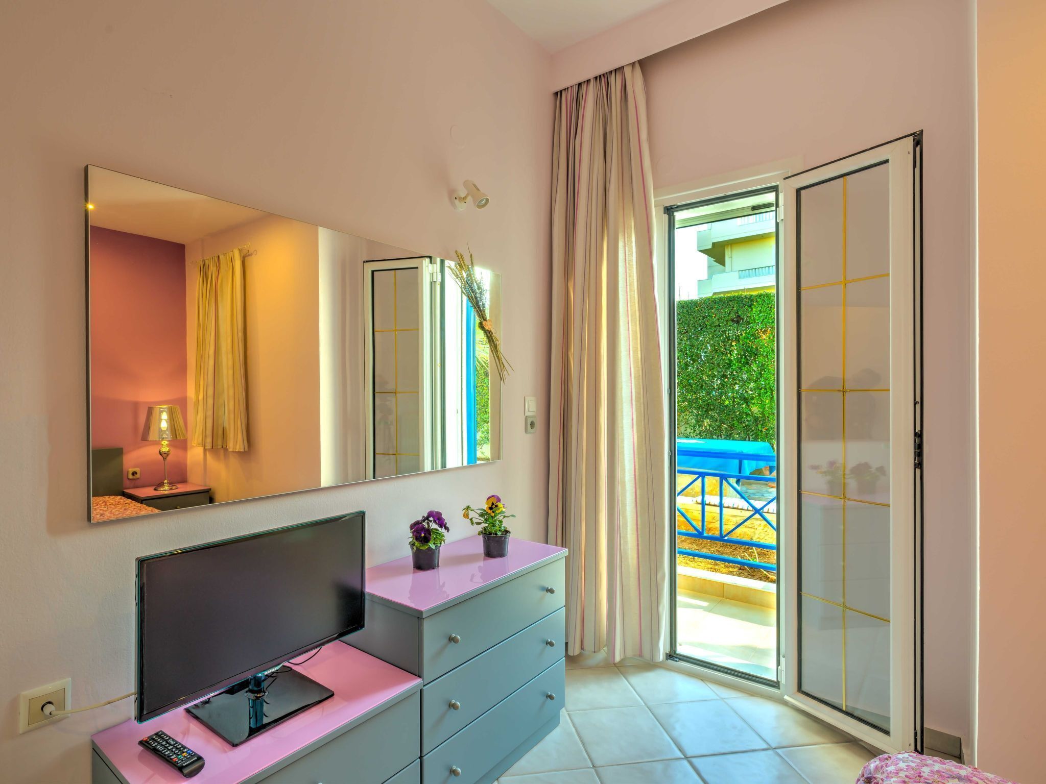 Une chambre à coucher comprenant un lit, une commode, un téléviseur, un miroir et une porte-fenêtre coulissante donnant sur un balcon.