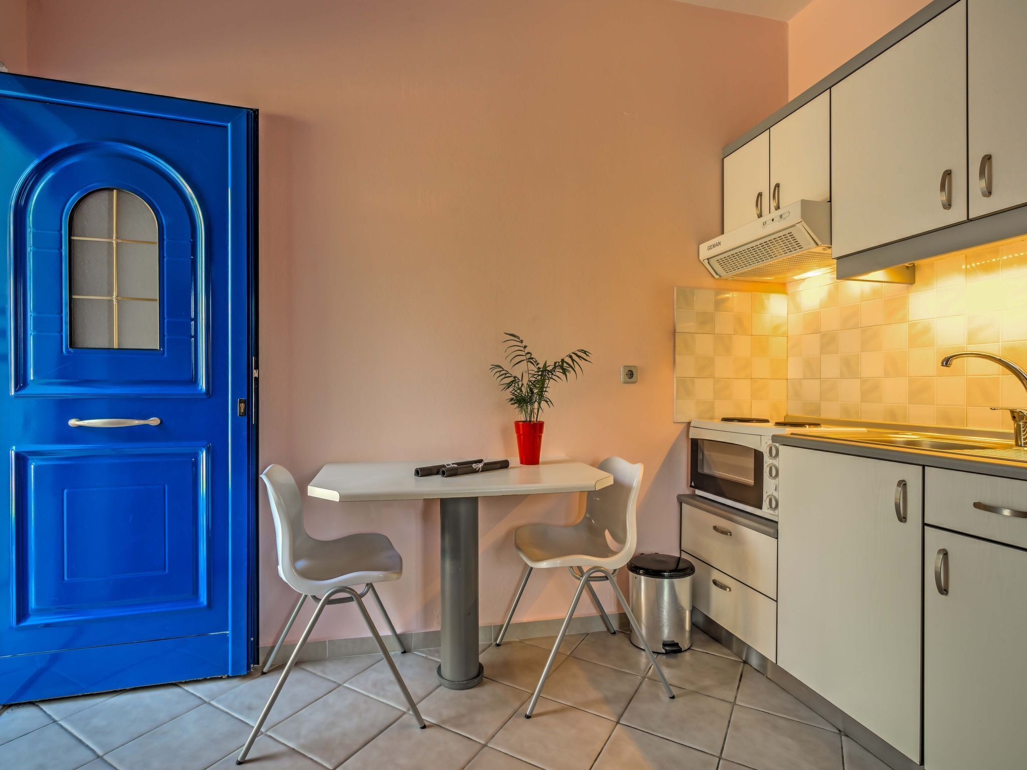 Una cocina funcional con puerta azul, una pequeña mesa de comedor, sillas, una planta en maceta y electrodomésticos modernos.