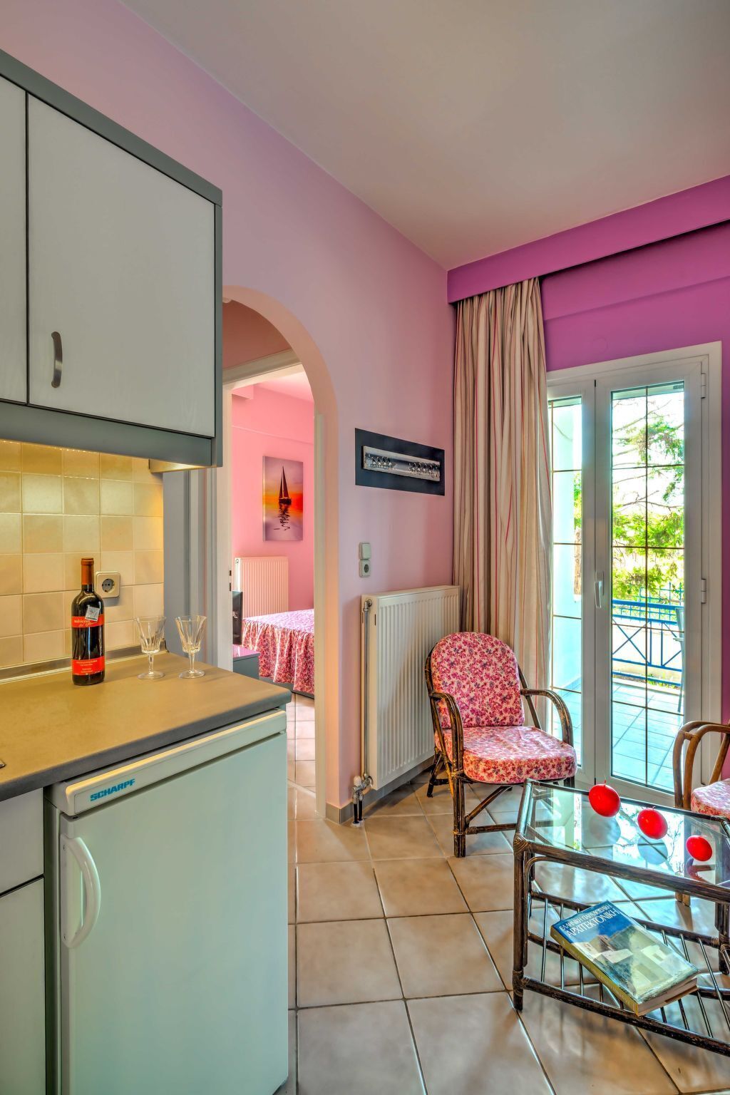 Una cocina con un refrigerador, una botella de vino y vasos sobre la encimera. Hay una silla y una mesa decorada con globos. Se ve una ventana con cortinas.