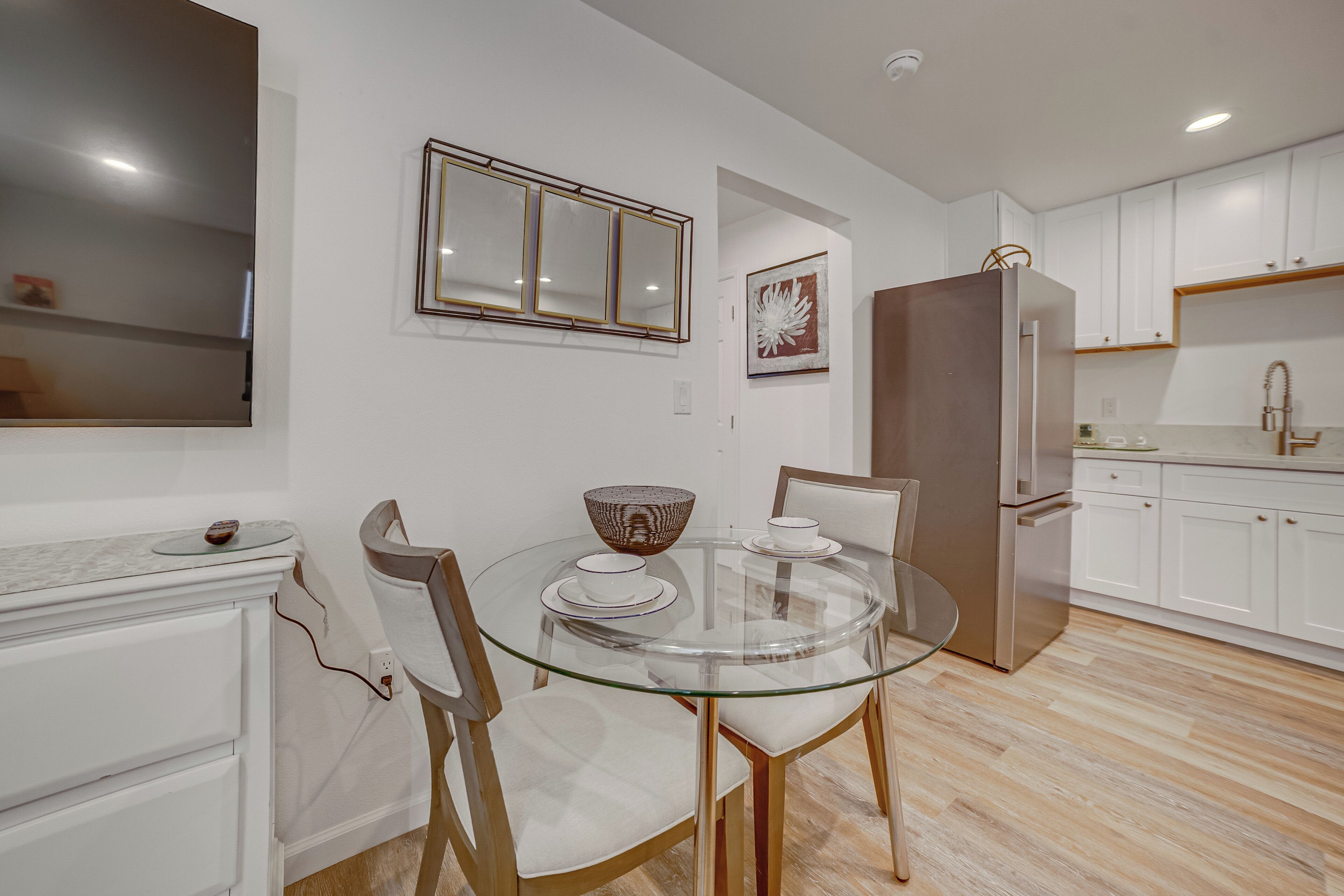 A modern kitchen with white cabinets, a stainless steel refrigerator, and a glass dining table with chairs.