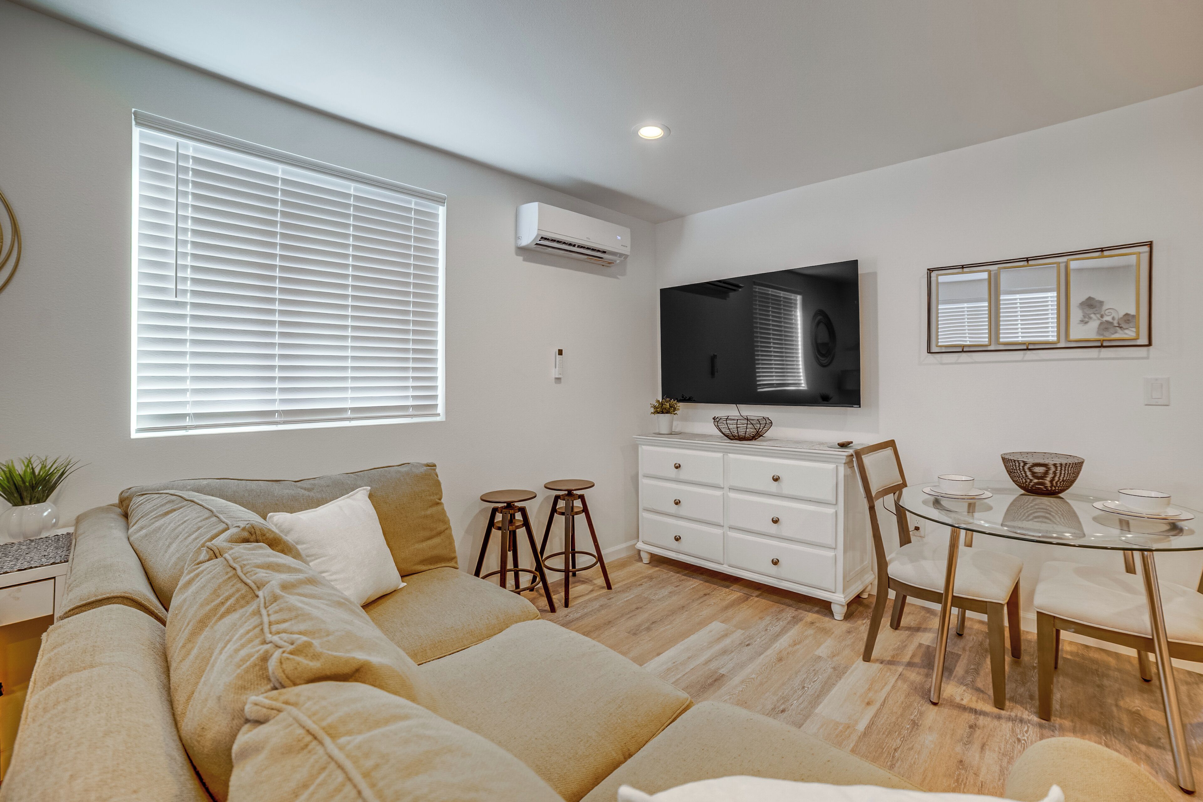 A modern living room with a sofa, a dining table with chairs, a flat-screen TV mounted on the wall, and a window with blinds.