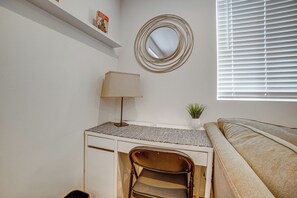 Ein Schlafzimmer mit einem Bett, einem Schreibtisch mit Stuhl, einer Lampe, einem Spiegel und einem Fenster mit Jalousien.