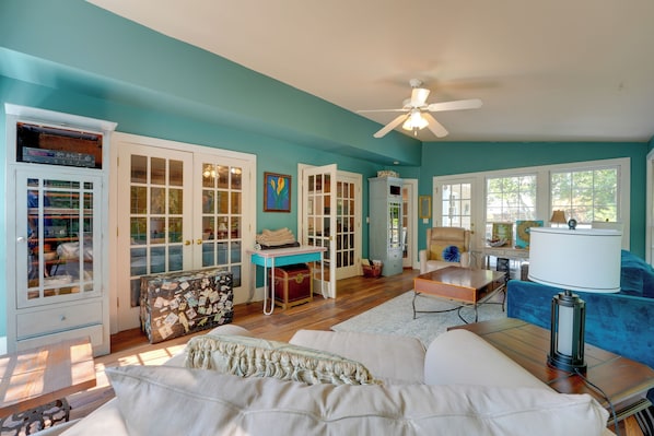 A room with teal walls, a ceiling fan, a desk with a lamp, a blue sofa, and a white armchair. - Group Getaway w/ Private Yard in Marietta (Marietta)