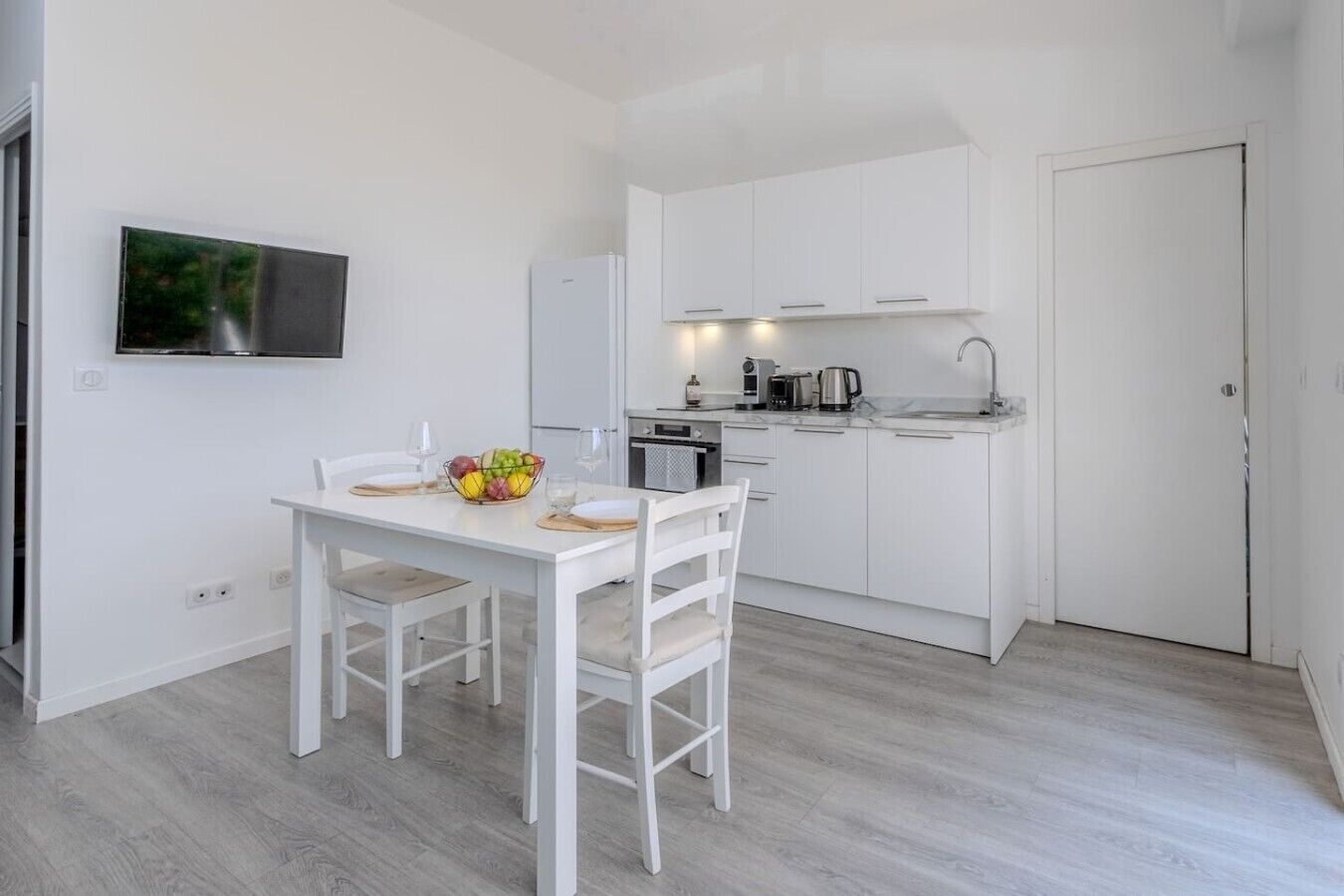 A compact kitchen with white cabinetry, a dining table with chairs, and a fruit bowl on the table.