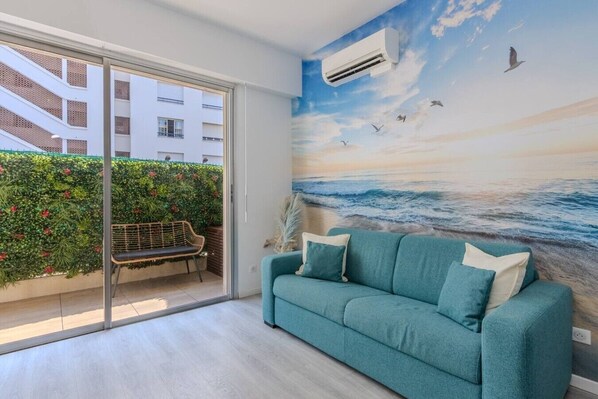 A room with a teal sofa, a bench, a mural of a beach scene, and a sliding glass door leading to an outdoor area with greenery. - Maupassant Terrace | 2 min from the beach (Antibes)