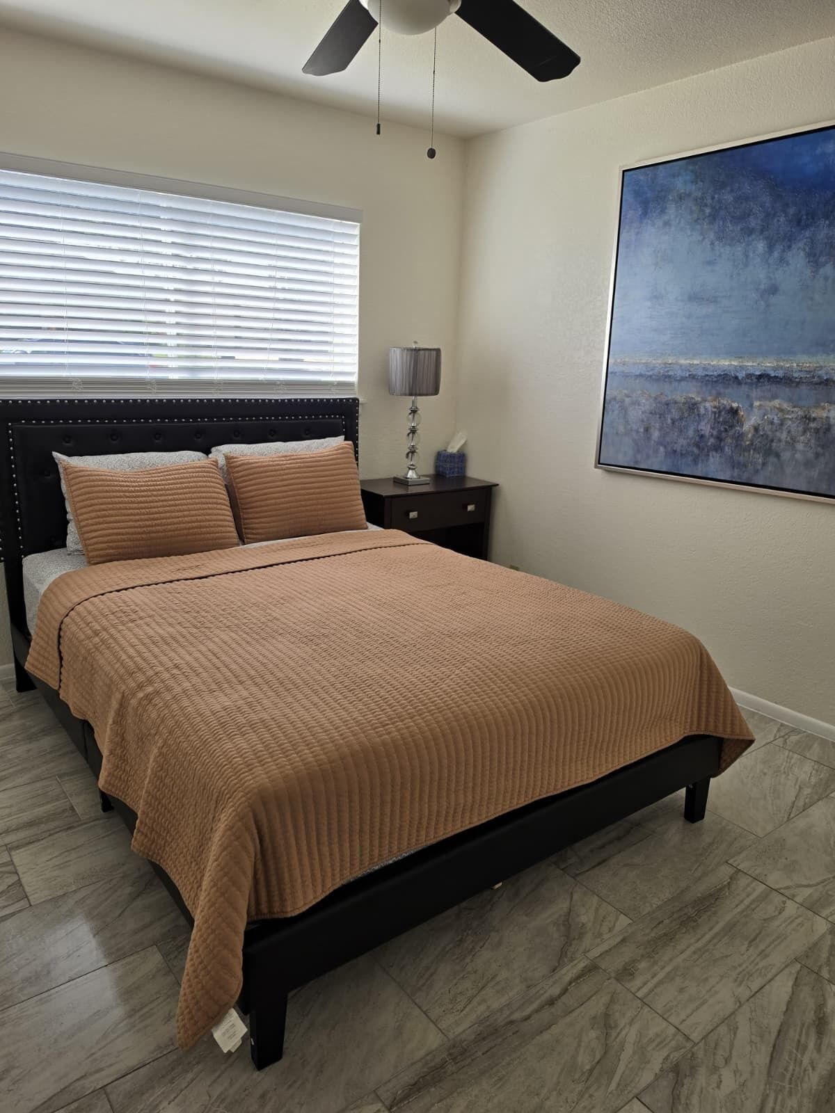 A bedroom with a bed, a nightstand, a lamp, a ceiling fan, and a painting on the wall.