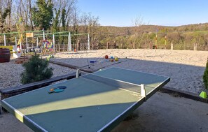 Ein Tischtennis-Tisch in einer sandigen Außenanlage, im Hintergrund ein Spielplatz.
